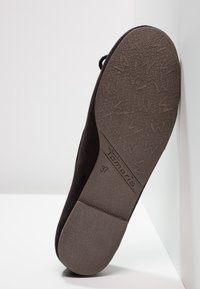 Semelle de chaussure plate noire avec grip texturé, marque « Tamaris », taille 37, reposant sur le talon contre des murs blancs.