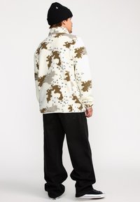 Veste en polaire blanche avec un motif abstrait marron et gris, présentant un col montant et une coupe décontractée ; assortie à un pantalon ample noir.