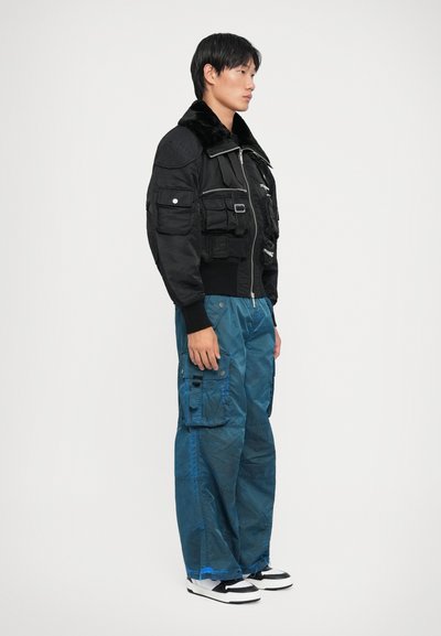 Chaqueta bomber negra con cuello de piel, múltiples bolsillos, cierre de cremallera, combinada con pantalones cargo azules holgados y zapatillas negras y blancas.