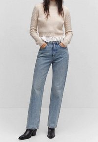 Pull beige côtelé avec un sous-couche blanche à volants, associé à un jean droit bleu clair et des bottines à talons noires.