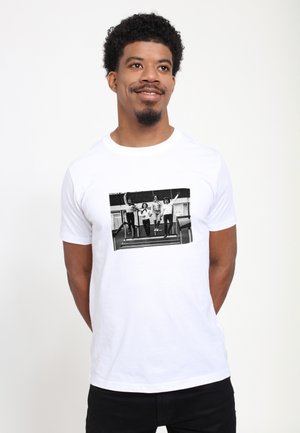 Mann trägt weißes T-Shirt mit schwarz-weißem Foto von vier Personen, die auf einer Treppe springen und freudig posieren.