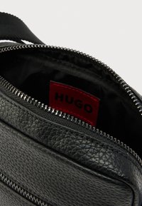 Mustast nahast ristitasku, millel on tekstureeritud pind, varustatud pealt lukuga ja punase sisemärgisega, millel on kiri "HUGO." Lukudetail on nähtav.