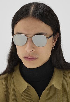 Persona con capelli lunghi e scuri indossa occhiali da sole aviator riflettenti, piccolo orecchino a cerchio, dolcevita nero e maglione a coste verde oliva.