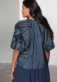 Kortærmet, mørkeblå top med tekstureret stof, der har et sort abstrakt mønster, rynket ryg og en afslappet pasform.