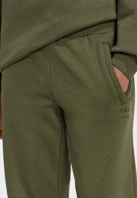 Zeleni pamukasti jogger hlače s mekom teksturom, koje imaju bočne džepove i reljefni logo na butu. Lagani i elastični pojas.