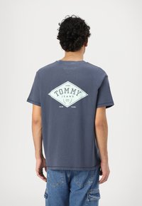Camiseta de algodón gris con un logo de diamante y el texto "TOMMY JEANS" en la parte posterior. Manga corta, cuello redondo y corte relajado.