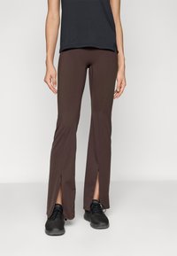 Brune treningsbukser med siden slisser nederst, laget av et elastisk materiale, sammen med svarte teksturerte sneakers og en tettsittende svart topp.