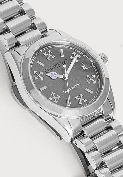 Montre-bracelet en métal argenté avec cadran gris, logo Off-White, aiguilles des heures et des minutes en forme de flèche, et texte "JUST WATCH" sur le cadran.