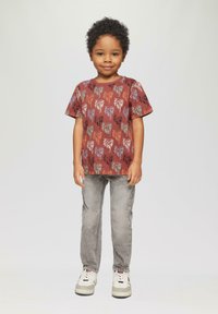 Brązowa koszulka z krótkim rękawem w wzór liści palmowych, zestawiona z jasnoszarymi dżinsami i białymi trampkami. Gładki materiał, luźny krój.