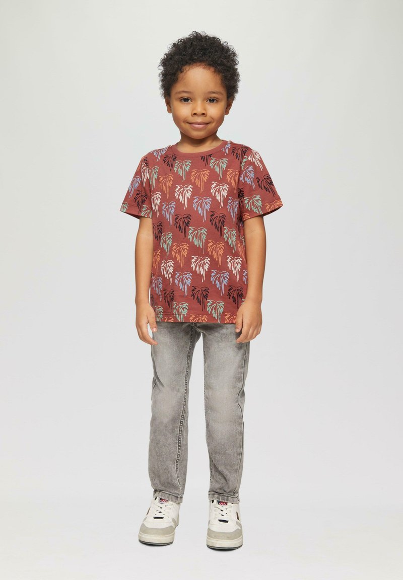 Brązowa koszulka z krótkim rękawem w wzór liści palmowych, zestawiona z jasnoszarymi dżinsami i białymi trampkami. Gładki materiał, luźny krój.