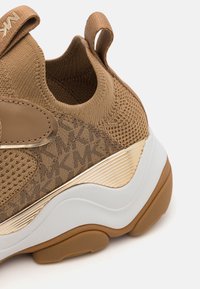 Shoe with breathable beige knit upper, patterned accents, and a white midsole featuring a gold metallic detail on the side.