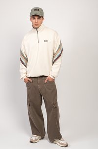 Felpa crema con zip e strisce colorate in teal, marrone e ruggine. Abbinata a pantaloni cargo marroni e sneakers beige. Accessorio: cappellino grigio.