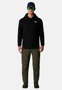 Pullover in pile nero con tasca a marsupio, abbinato a pantaloni cargo verde scuro con tasche con zip e scarponi da trekking neri.