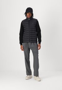Black puffer vest with horizontal stitching, paired with a black long-sleeve shirt, grey trousers, and beige shoes. Black cap with logo.