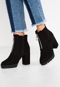 Botas de ante negras hasta el tobillo con un tacón grueso, cremalleras laterales y suelas texturizadas, combinadas con unos jeans de mezclilla claros y cortos.