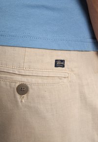 Beige linnen shorts met een achterzak, voorzien van een knoopsluiting en een blauwe geweven label met de tekst "Petrol". Soepele textuur, casual ontwerp.