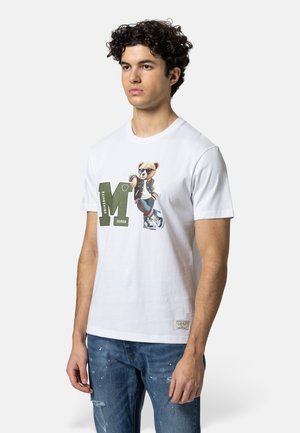 Jonge man draagt een wit T-shirt met een grote groene "M" en een beer met zonnebril en varsityjack die erop leunt, gecombineerd met een blauwe spijkerbroek.