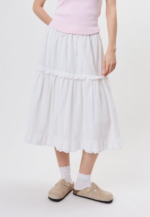 Femme portant une jupe midi blanche à étages, un haut rose sans manches, des chaussettes blanches à volants et des sabots beiges à enfiler, debout devant un fond uni.