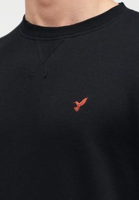 Μαύρο φούτερ με στρογγυλή λαιμόκοψη, υφασμάτινες ραφές με υφή και μικρό κόκκινο λογότυπο πουλιού κεντημένο στο στήθος.