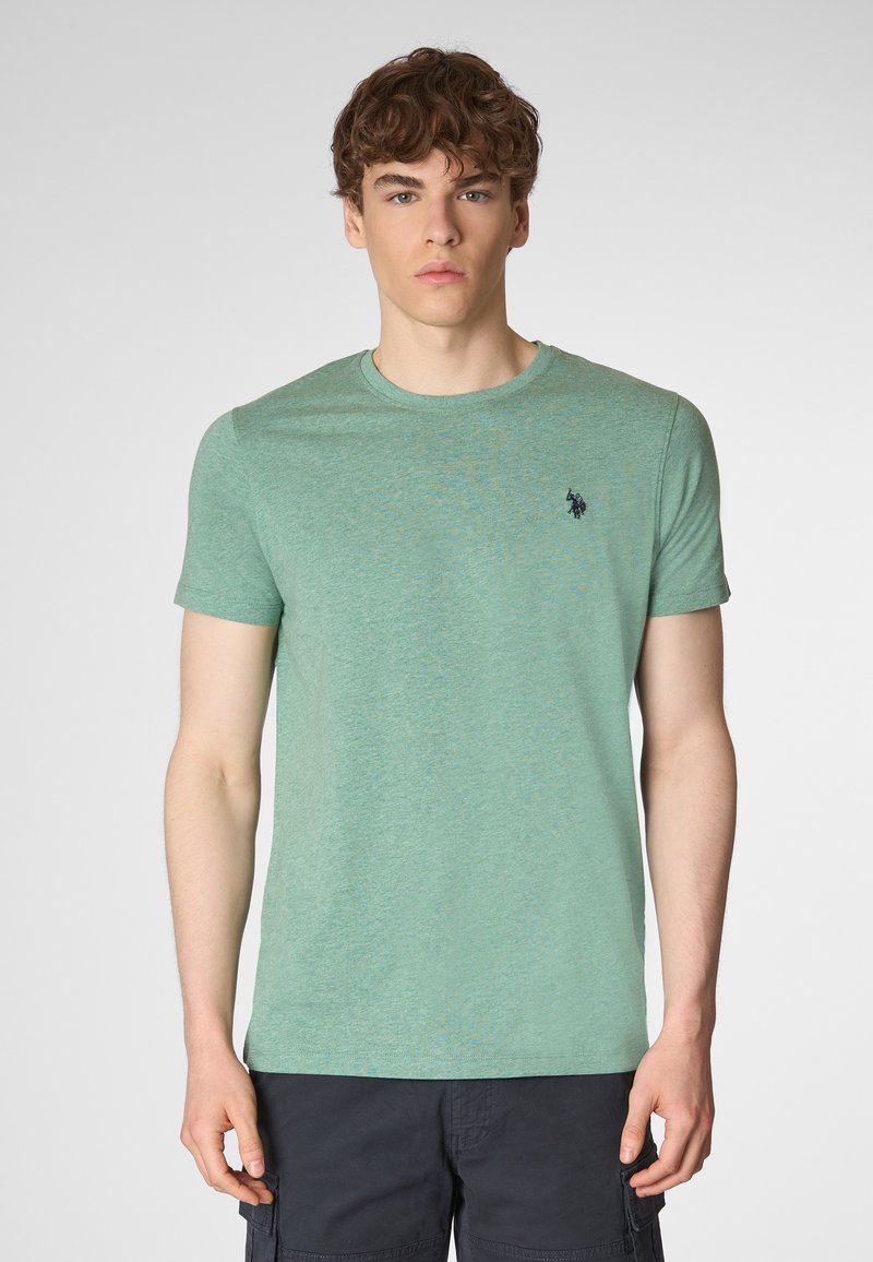 Un giovane uomo è in piedi contro uno sfondo chiaro, indossando una maglietta verde con un piccolo logo, guardando direttamente verso la fotocamera.