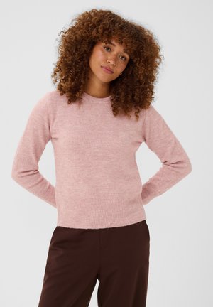 Kvinde med krøllet hår iført en lyserød sweater og mørkebrune bukser, stående med hænderne bag ryggen foran en ensfarvet baggrund.