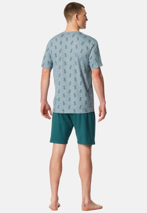 Mand står barfodet med ryggen til iført lyseblå T-shirt med mønster af mørkeblå ananas og mørkegrønne shorts.