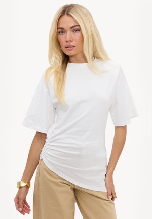 Femme blonde portant un haut blanc froncé à manches courtes et un pantalon beige, accessoirisée d’un bracelet et d’une bague en or.