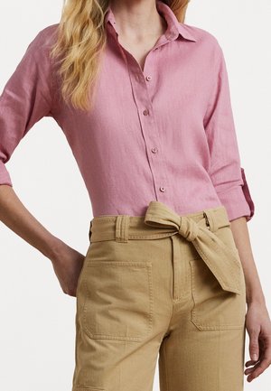 Frau trägt ein pinkfarbenes Hemd mit hochgekrempelten Ärmeln und beige, hoch taillierte Hosen, die mit einem Stoffgürtel in einer Schleife gebunden sind.