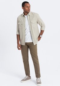Chemise boutonnée gris clair avec deux poches poitrine sur un T-shirt blanc, associée à un pantalon vert olive et des baskets grises.
