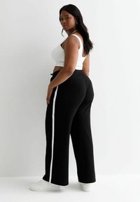 Vrouw in een witte crop top, zwarte wijde broek met witte zijstrepen, en witte sneakers, staand tegen een effen witte achtergrond.