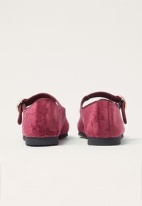 Zapatillas rojas sin cordones con un acabado texturizado, que cuentan con correas elásticas y un acento en forma de anillo dorado en la parte posterior. Suelas negras de perfil bajo.