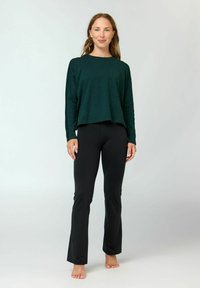 Frau steht barfuß und trägt ein dunkelgrünes Langarmshirt sowie schwarze Schlaghosen vor hellem, einfachem Hintergrund.