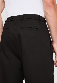 Pantalons noirs avec une texture lisse, une taille élastique, deux poches arrière et une étiquette logo noire sur la poche droite.