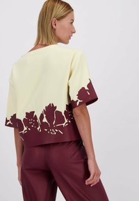 Vrouw met kort blond haar, gekleed in een crèmekleurig top met een donker bloemmotief aan de rand en bijpassende donker kastanjebruine broek, gezien van achteren.