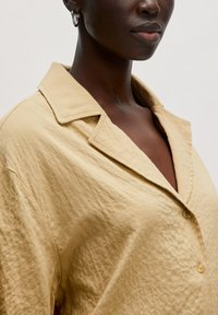 Beige, strukturiertes Hemd mit Kragen und tiefem V-Ausschnitt. Verfügt über eine lockere Passform und dezente Falten für einen lässigen Look.