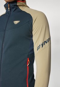 A navy and beige jacket featuring a zippered front, textured fabric, orange zipper accents, and a logo on the sleeve.