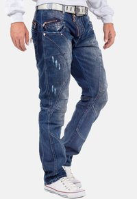 Jeans in denim blu con molte tasche con zip, dettagli consumati e cuciture a contrasto. Indossati con sneakers bianche e un maglione grigio.