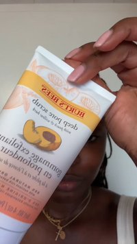 En person som holder en tube med Burt’s Bees dyp pore-peeling med fersken og pilbark, iført en hvit topp og gullkjeder.