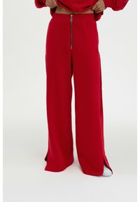 Rote weit geschnittene Sweatpants mit einem Kordelzugbund, seitlichen Reißverschlusstaschen und hohen Schlitzen an den Seiten. Weicher Stoff.