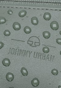 Superficie verde texturizzata con motivi circolari in rilievo e gocce; presenta il nome del marchio in rilievo "JOHNNY URBAN" sotto un'icona di faccina sorridente.