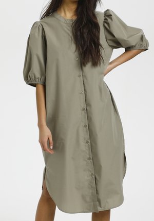 Woman wearing a loose, buttoned olive-green dress with puffed elbow-length sleeves and a rounded neckline.