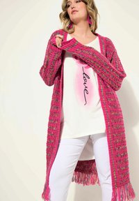 Długi kardigan z teksturowanej dzianiny w jasnoróżowym kolorze z wielokolorowymi akcentami, z frędzlami na dole. Pod spodem biała koszula.