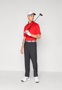 Tricou polo roșu, pantaloni de golf negri, șapcă albă și pantofi de golf albi; ținând un club de golf; caracteristicile includ un buzunar pe tricou și mănuși.