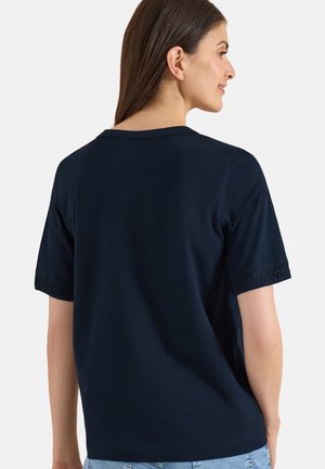 Donna con lunghi capelli castani che indossa una maglietta tinta unita blu scuro a maniche corte con bordo in pizzo sui bordi delle maniche, vista di lato e leggermente girata indietro.