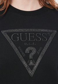 Sudadera negra con logo triangular brillante de Guess, que incluye "GUESS U.S.A.", un signo de interrogación y las palabras "WASHED JEANS" en la parte delantera.