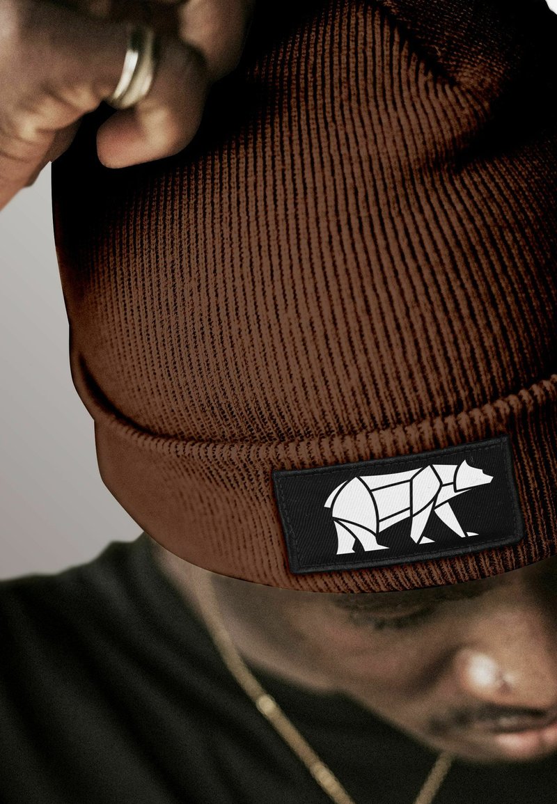 Person wearing ribbed brown beanie with a geometric white bear logo patch, adjusting it with a hand wearing a silver ring.