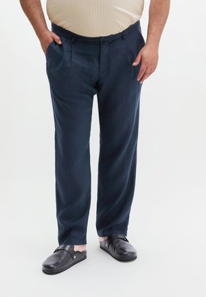 Mann trägt marineblaue Hose und schwarze Slipper, steht mit den Händen in den Taschen vor einem einfarbigen Hintergrund.