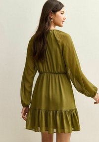 Vestido verde oliva con mangas largas, dobladillo con volantes y textura plisada. Presenta una cintura ajustada y una silueta suelta y fluida.