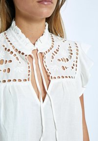 Bílá blůza s řaseným límcem, obsahující jemné vyřezávané vzory a šňůrky u výstřihu. Měkká tkanina s jemnou strukturou.