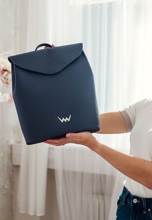 Person holding a dark blue backpack with a quilted flap and silver logo in a bright room with sheer curtains.